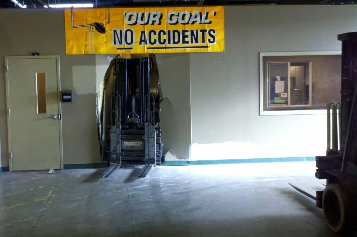 A forklift is visibly embedded in a wall, having crashed through it, directly beneath a yellow banner that reads 'OUR GOAL'…