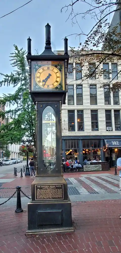 A tall, ornate black steam clock with a glowing clock face and Roman numerals stands on a brick sidewalk in a city, with a…