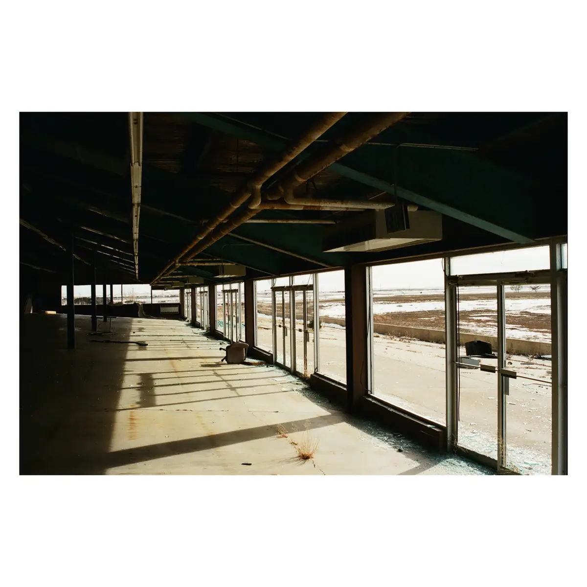 An abandoned, empty building with broken glass windows and exposed pipes, showing a desolate interior with sunlight…