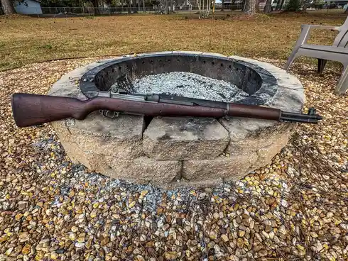 A wooden-stocked rifle with a metal barrel rests horizontally across the stone rim of an outdoor fire pit filled with ash…
