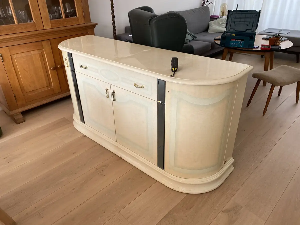 A cream-colored, ornate, curved cabinet with gold handles sits in a living room, with a tape measure resting on top. In the…