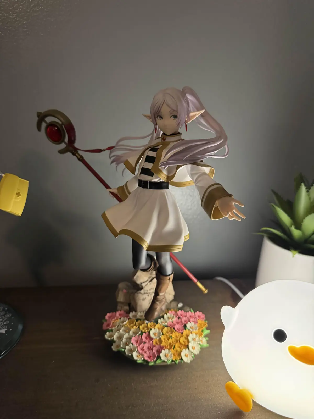 A figurine of an anime character with long lavender hair, pointed ears, and a white and gold outfit holding a red staff…