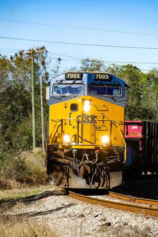 A yellow and blue CSX train engine, number 7003, is shown approaching on tracks with its headlights on under a clear blue…