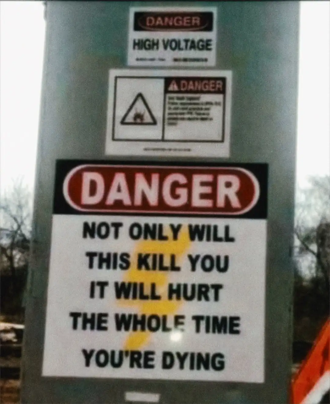A close-up of a utility pole covered in multiple warning signs, with the most prominent one reading 'DANGER NOT ONLY WILL…