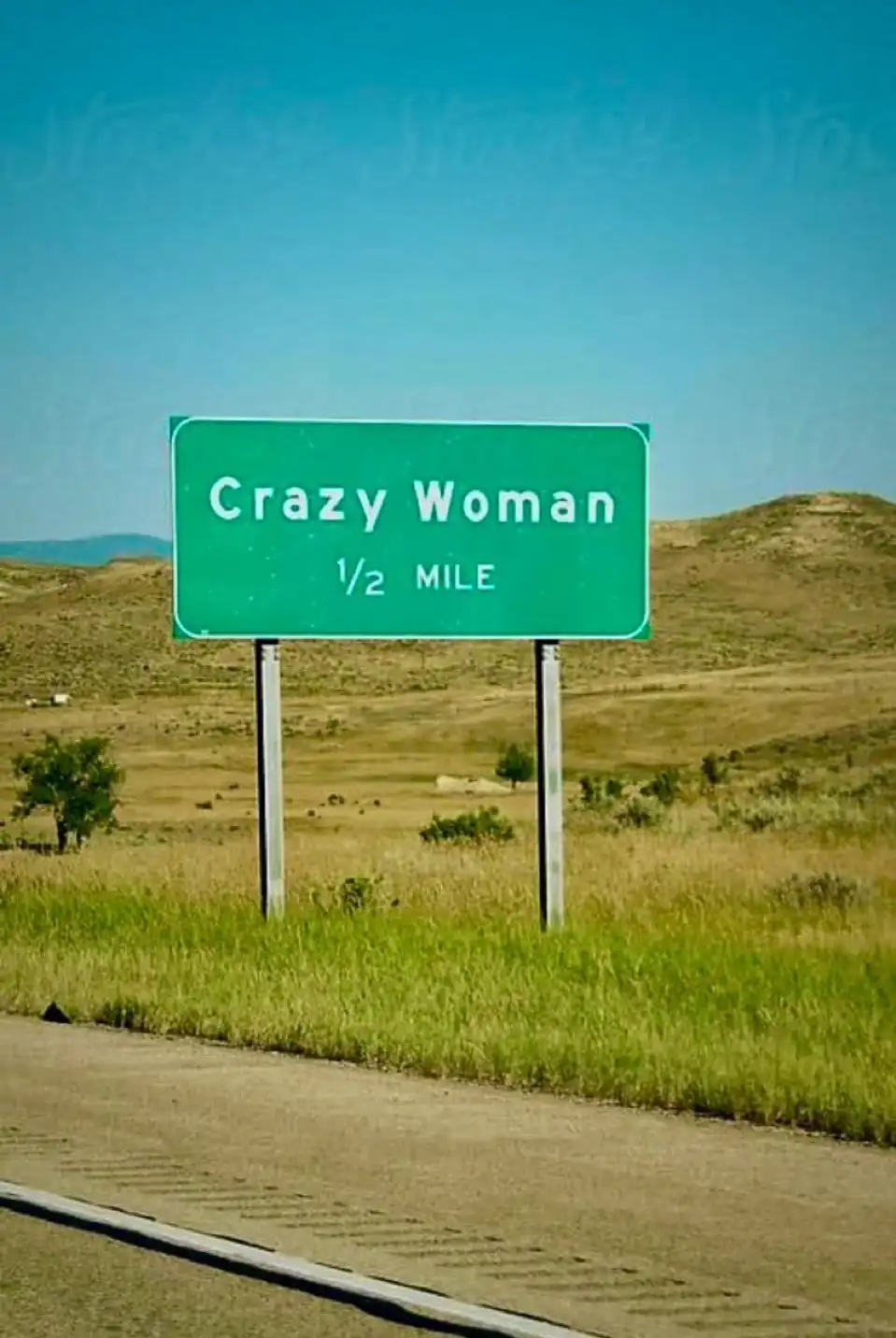 A green highway sign in a dry, grassy landscape under a clear blue sky reads 'Crazy Woman 1/2 MILE', humorously indicating a…
