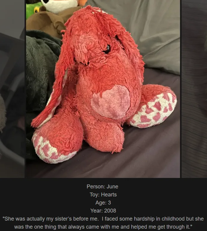 A heavily worn, frayed red plush bunny toy with long floppy ears and a pink heart on its chest sits on a dark fabric…