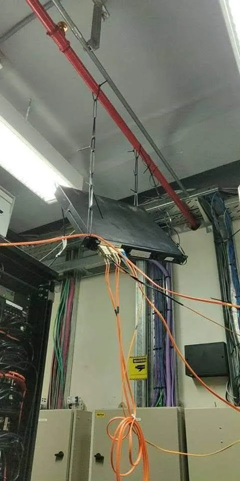 A laptop hangs suspended from a ceiling by a chain, surrounded by tangled orange cables and server equipment. The overlaid…