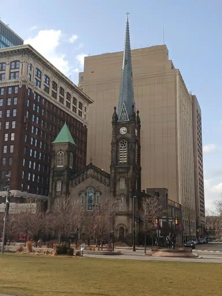 A photo of a historic stone church with a tall, pointed spire and stained glass windows, situated in a city surrounded by…