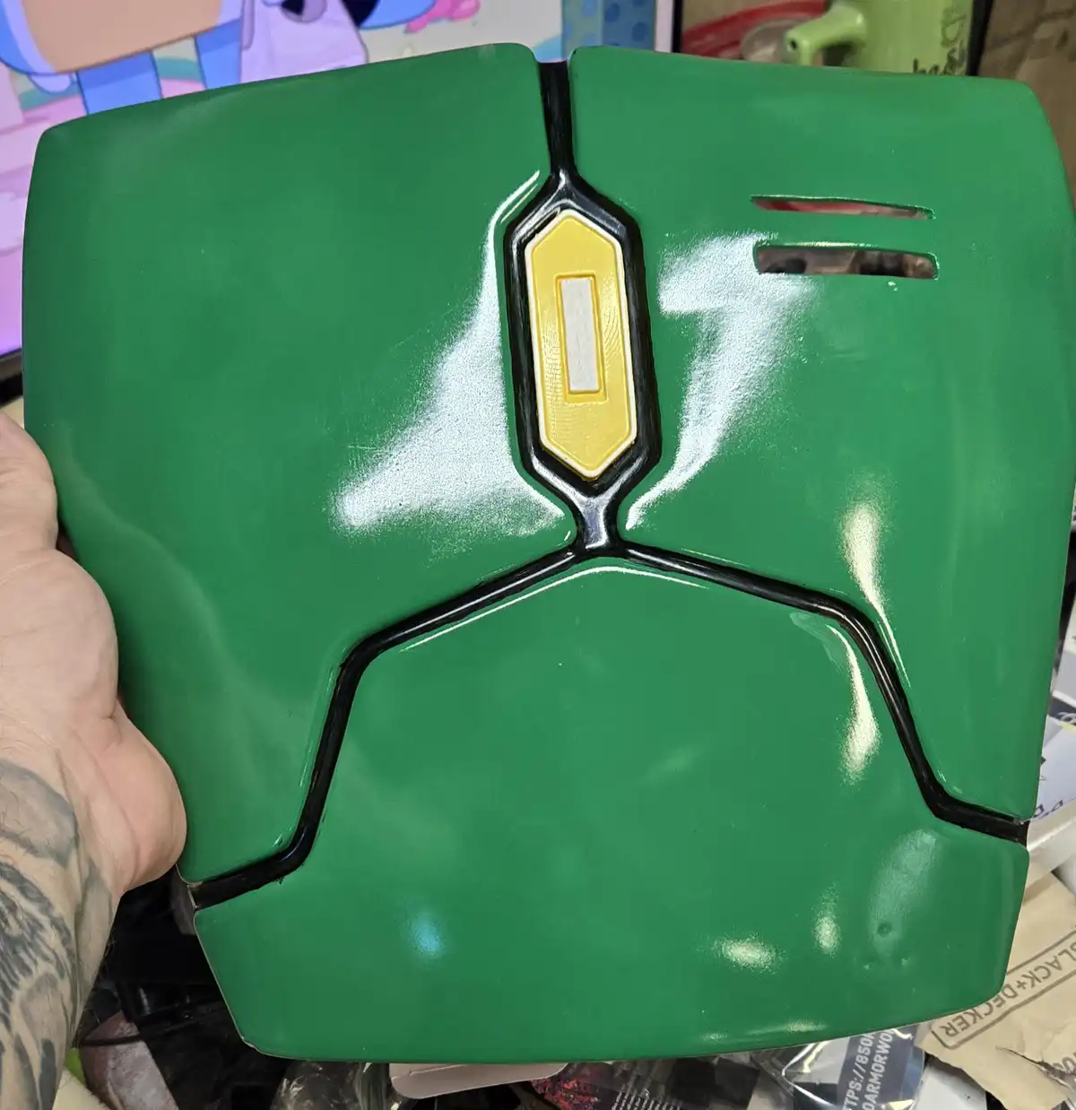 A hand holds a green, glossy, toaster-shaped object with black seams and a yellow rectangular emblem, resembling a Boba Fett…