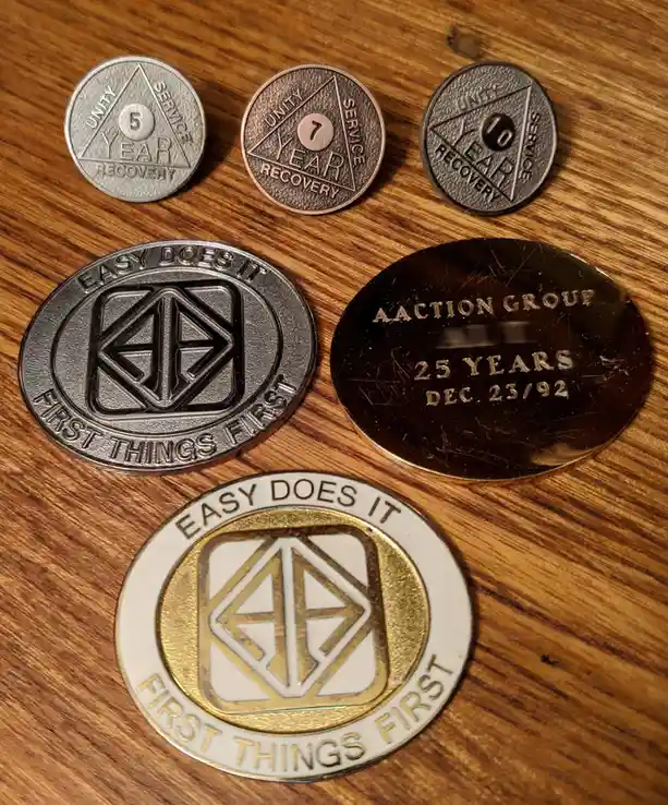 Six commemorative tokens lie on a wooden surface: three circular recovery milestone tokens (5, 7, and 10 years) and three…