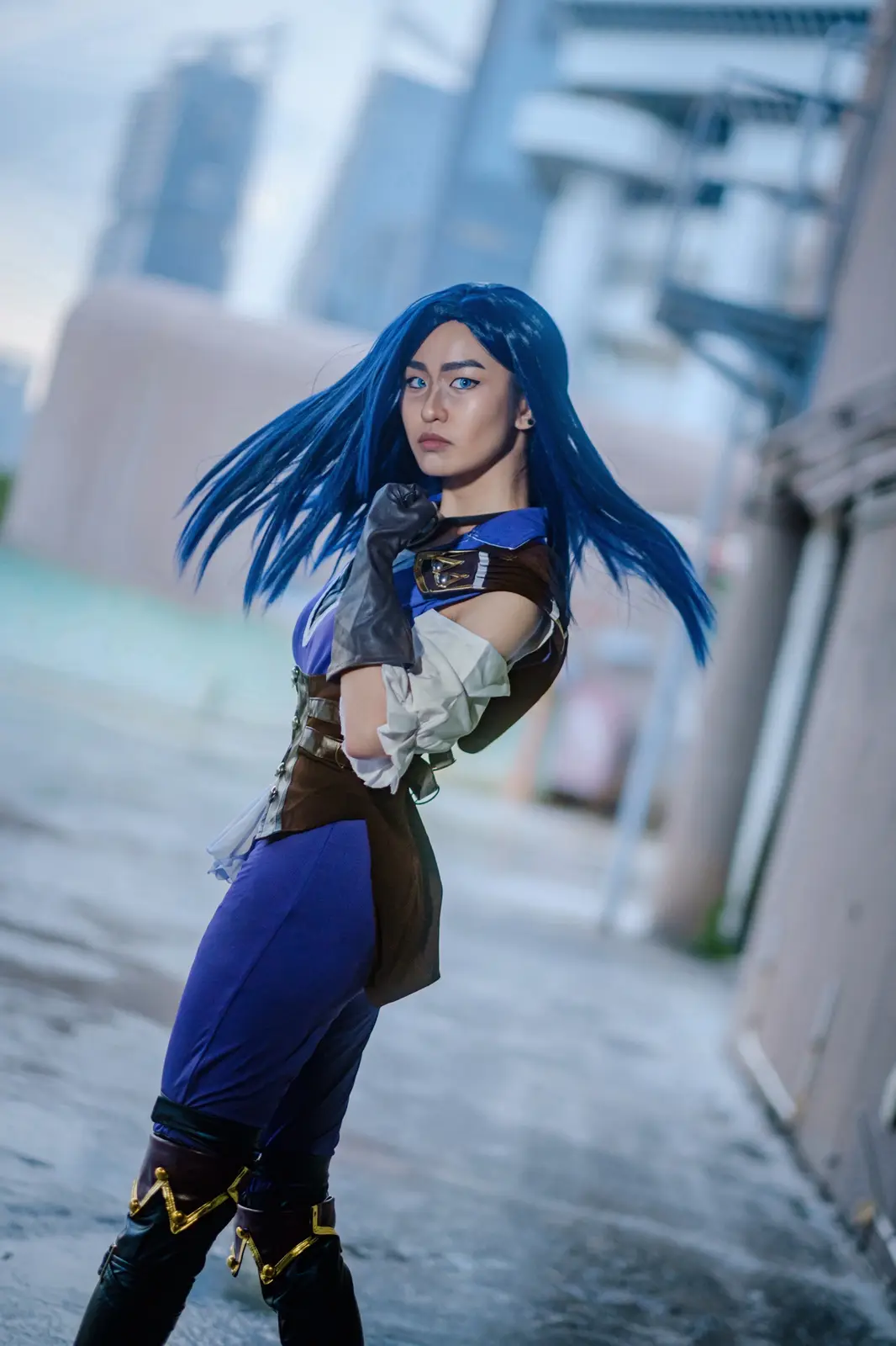 A person with long blue hair and blue eyes is cosplaying as a character in a blue, white, and brown outfit with black boots…