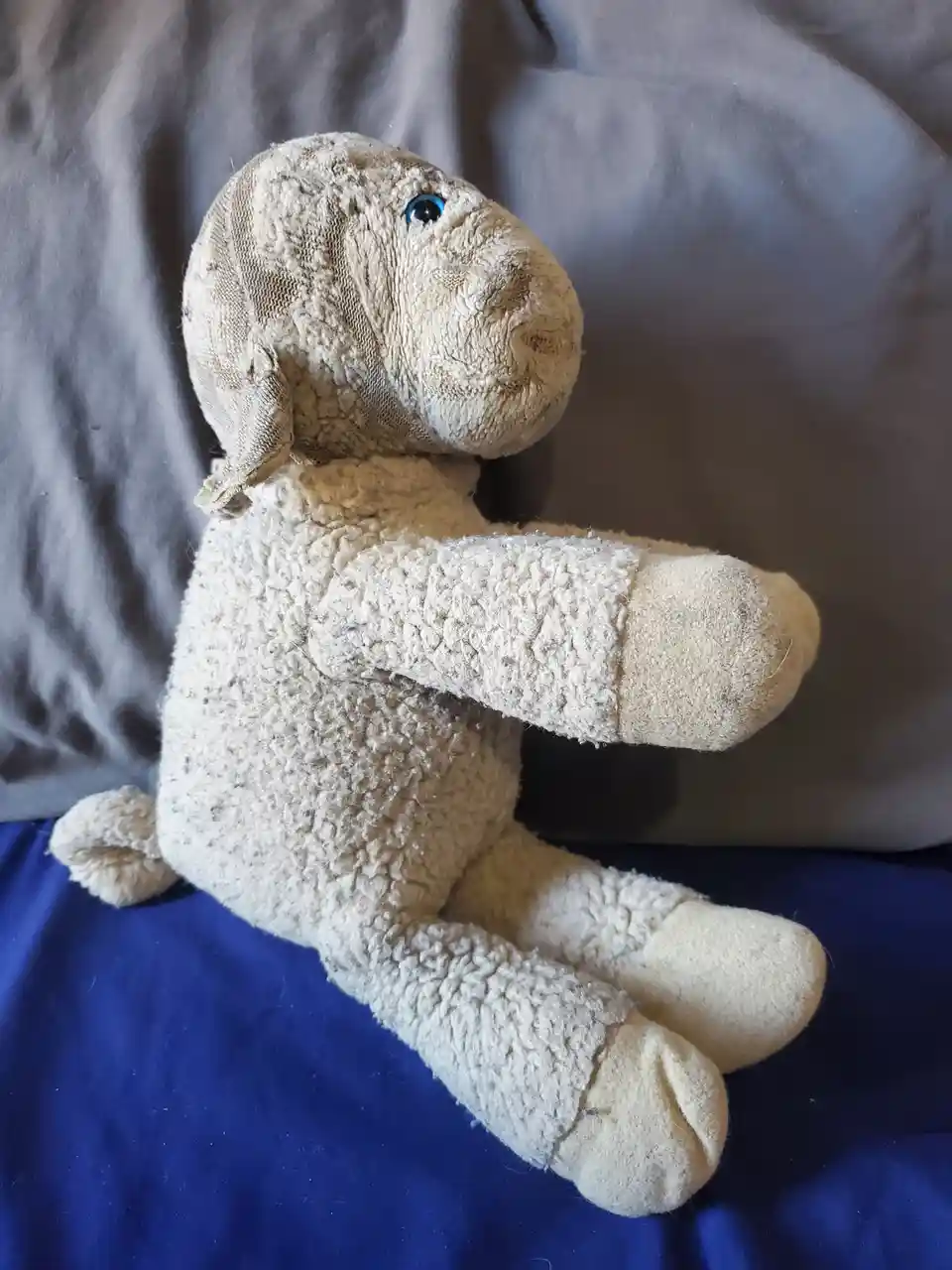 A very worn, beige stuffed sheep toy with one blue eye and frayed fabric sits on a blue surface against a gray background…
