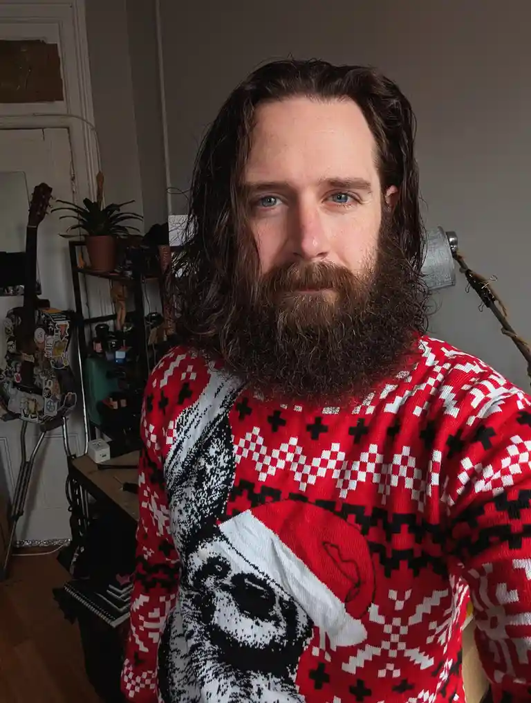 A man with long brown hair and a full beard stares directly at the camera while wearing a red, black, and white patterned…