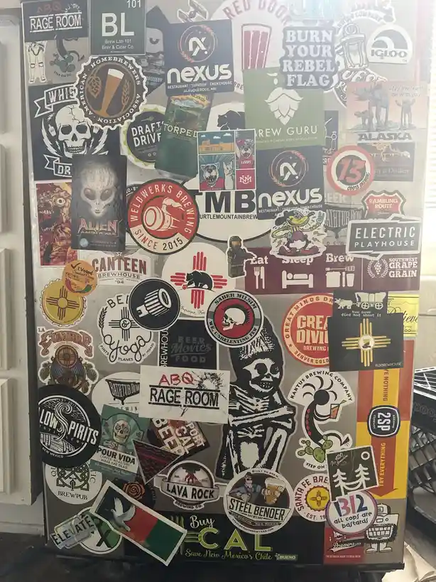 A refrigerator door is covered in a dense collage of beer, brewery, and local New Mexico-themed stickers, including logos…