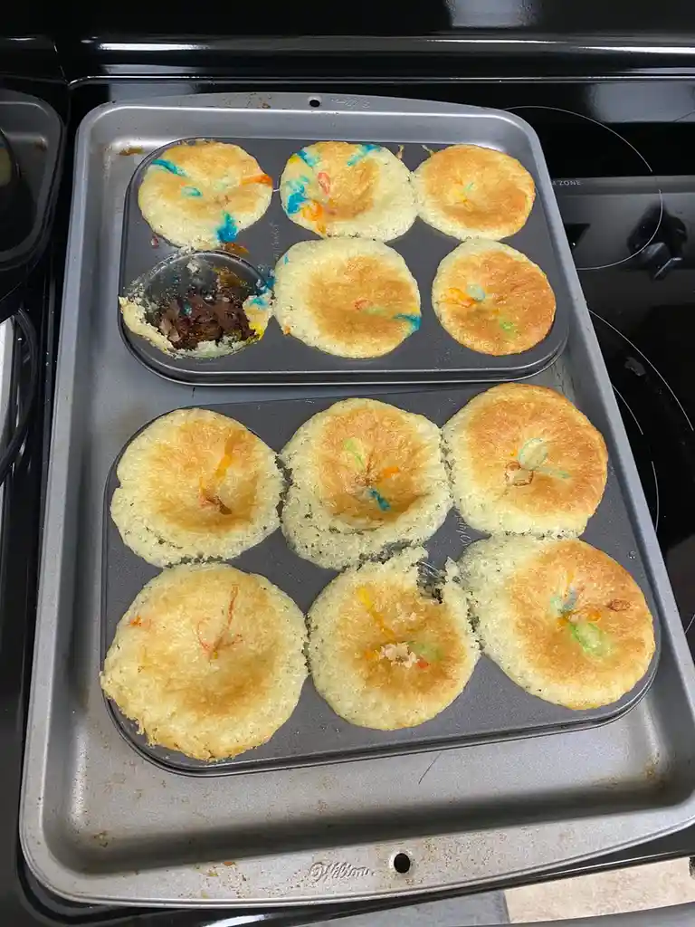 Two metal muffin tins sit on a baking sheet, each containing golden-brown baked goods with colorful sprinkles. One muffin in…