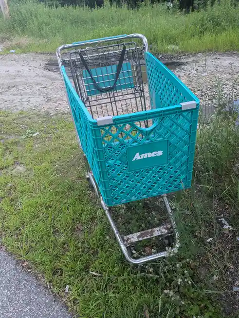 A teal Ames shopping cart with a lattice-style plastic basket and metal frame is abandoned in a grassy, muddy area near a…