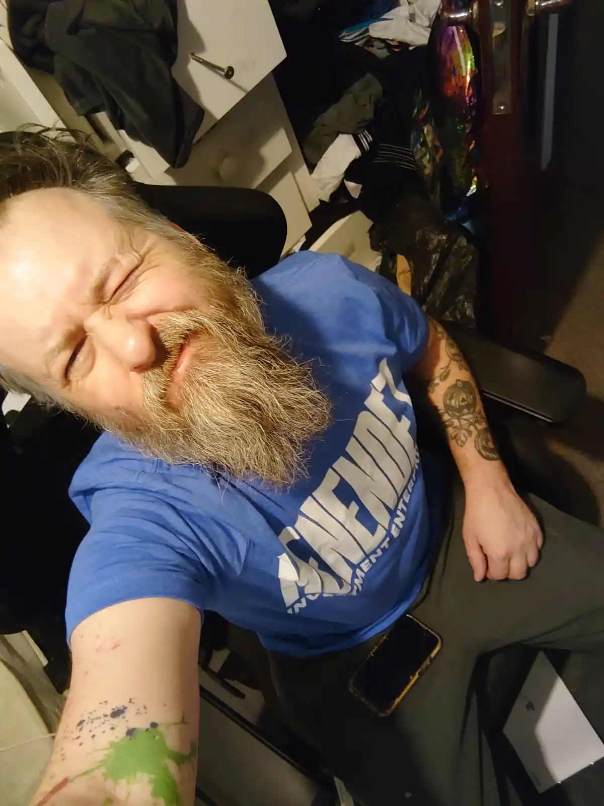 A man with a long beard and tattoos sits in a messy room, grimacing with his eyes squeezed shut. His left arm is covered in…
