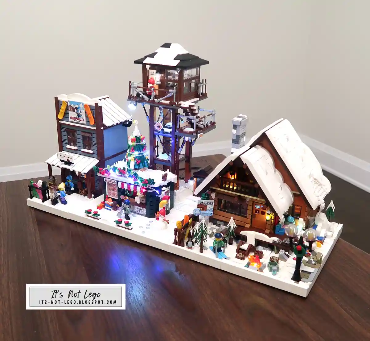 A detailed, illuminated LEGO winter village diorama featuring a ski lodge, a tower, a shop with a 'White Valley' sign, and…