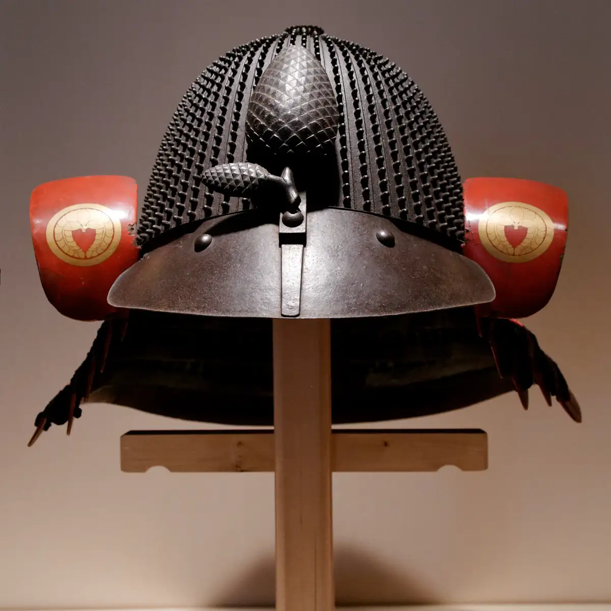 A close-up photograph of a traditional Japanese samurai helmet, or kabuto, displayed on a wooden stand. The helmet features…