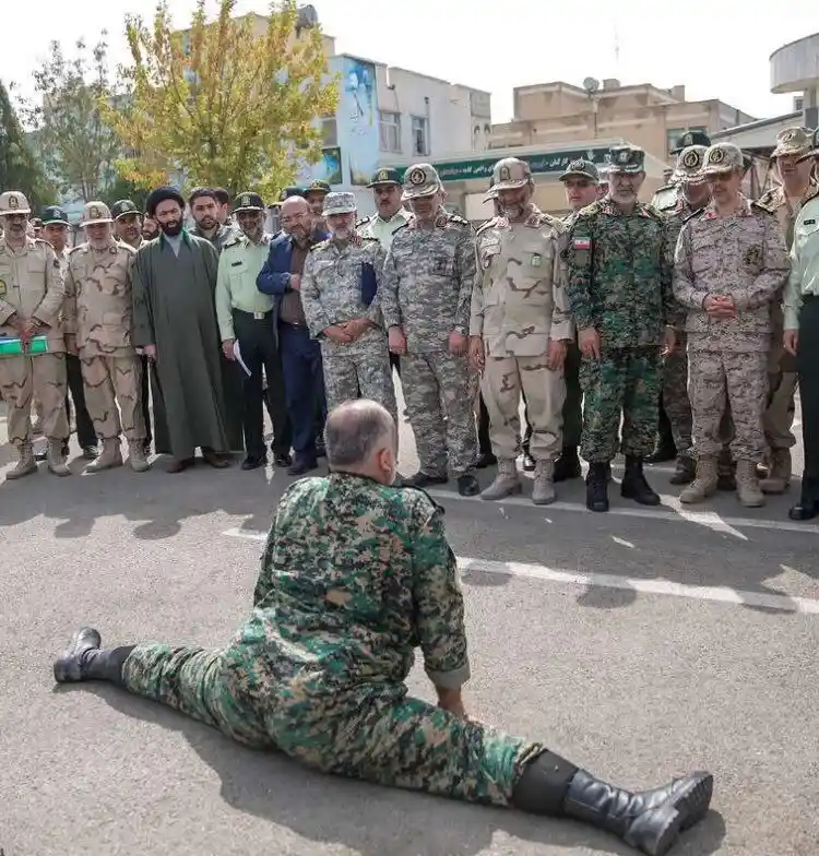 A man in a green military camouflage uniform performs a full split on an asphalt surface while facing a group of standing…
