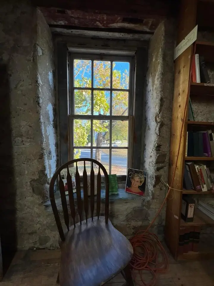 A wooden chair sits in front of a window in a rustic room with exposed stone walls and a bookshelf labeled 'Philosophy'…