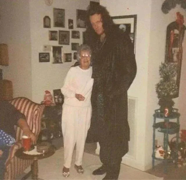 A grainy, vintage-style photo shows a very tall man in a dark, textured coat standing beside a much shorter elderly woman in…