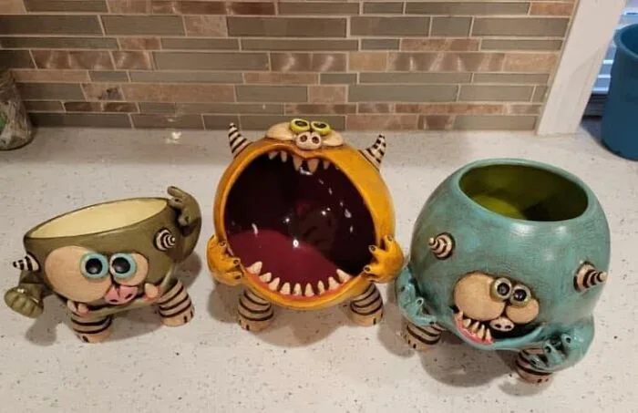 Three ceramic monster-themed bowls sit on a kitchen counter. The central yellow bowl has a wide-open mouth with teeth and a…