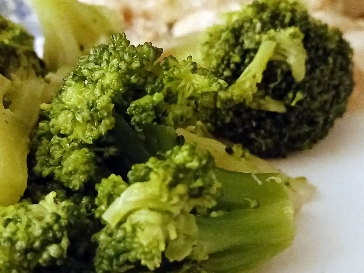 A close-up photo of cooked broccoli florets on a plate, with one floret in the background appearing dark and charred. The…