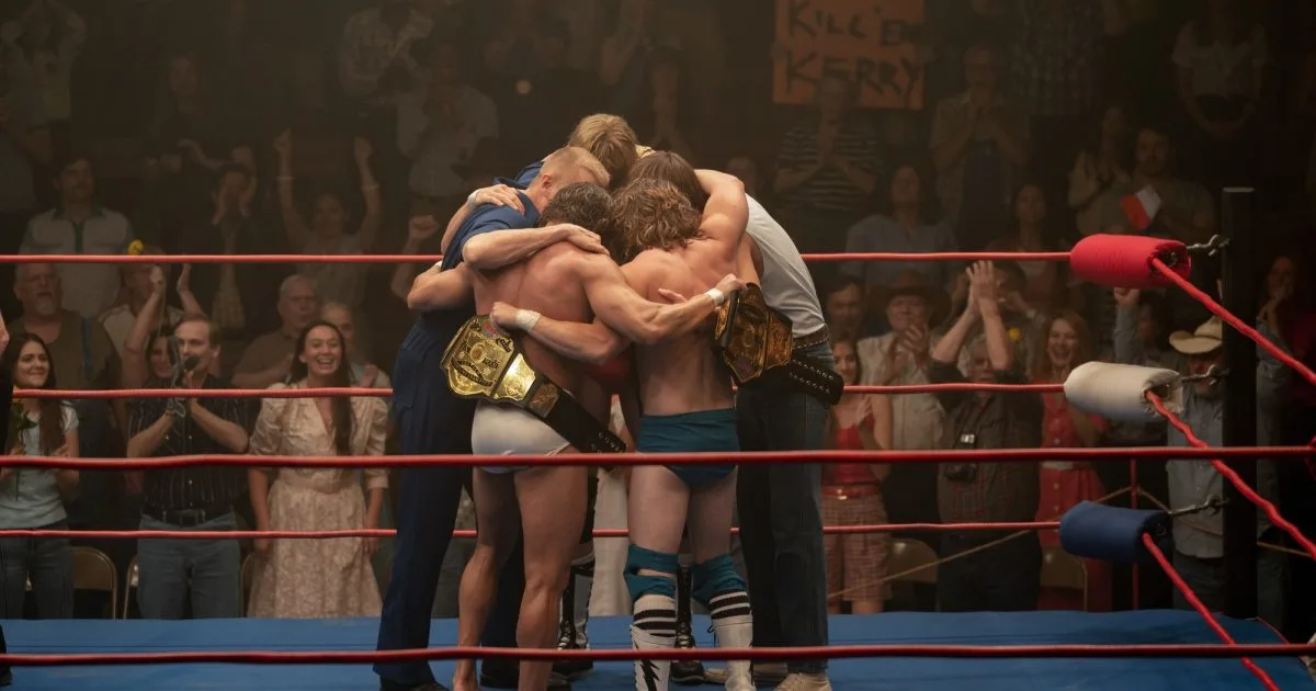 In a wrestling ring, two wrestlers in trunks and championship belts are embraced by two men in casual clothes, while a crowd…