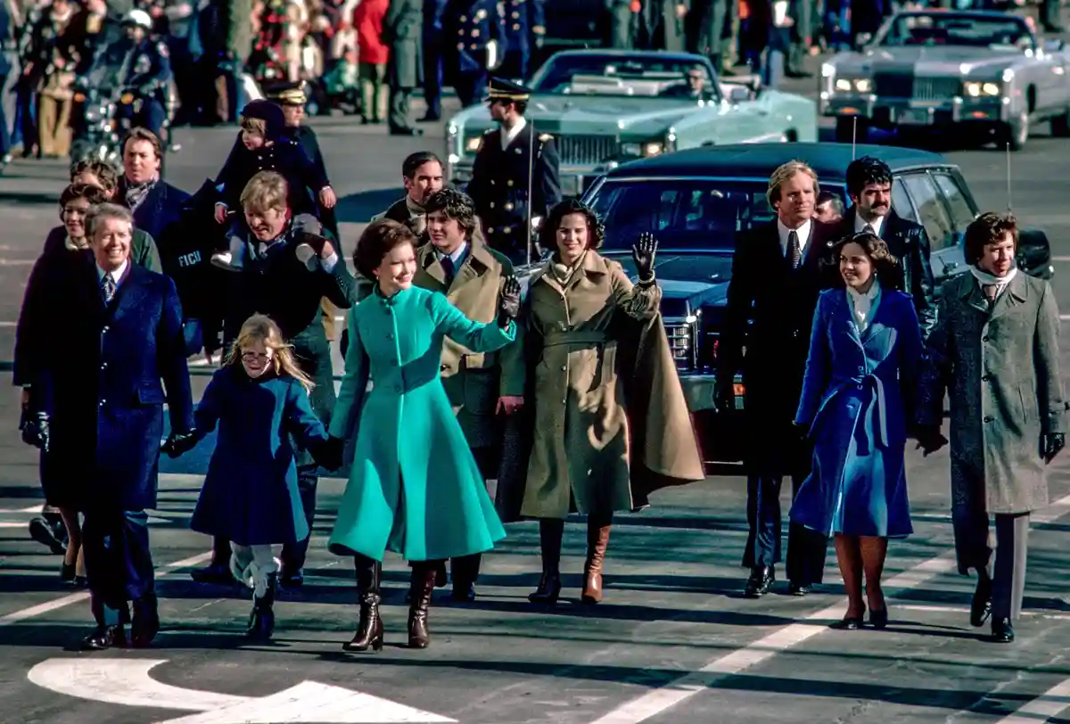 A historical photo shows the Carter family walking outdoors in winter coats, smiling and waving to crowds. The image…