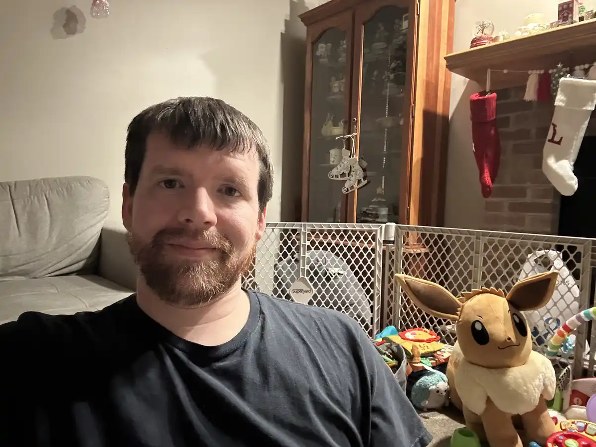 A man with a beard and dark t-shirt takes a selfie in a living room, smiling slightly. Behind him is a white baby gate, a…