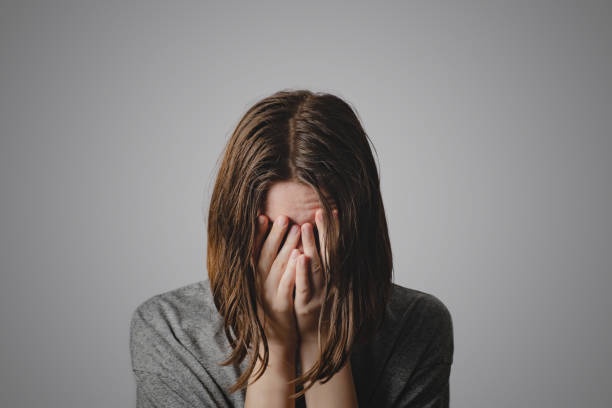 A person with brown hair, wearing a gray shirt, covers their face with both hands against a plain gray background. The…