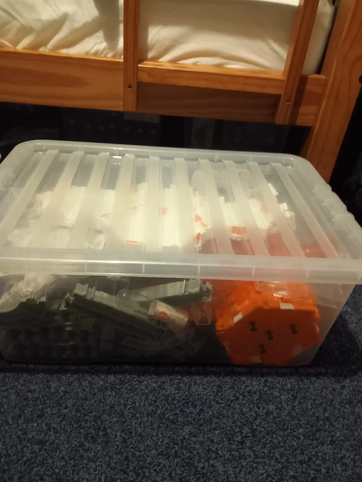 A clear plastic storage bin filled with LEGO pieces sits on a carpeted floor under a wooden bunk bed. The bin's lid is…