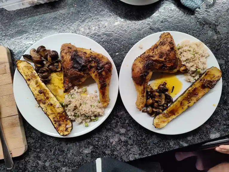Two identical plates of roasted chicken thighs, roasted zucchini, sautéed mushrooms, and rice with peas sit side-by-side on…