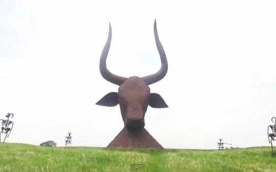 A large, rust-colored metal sculpture of a bull's head with prominent curved horns rises from a grassy field under an…