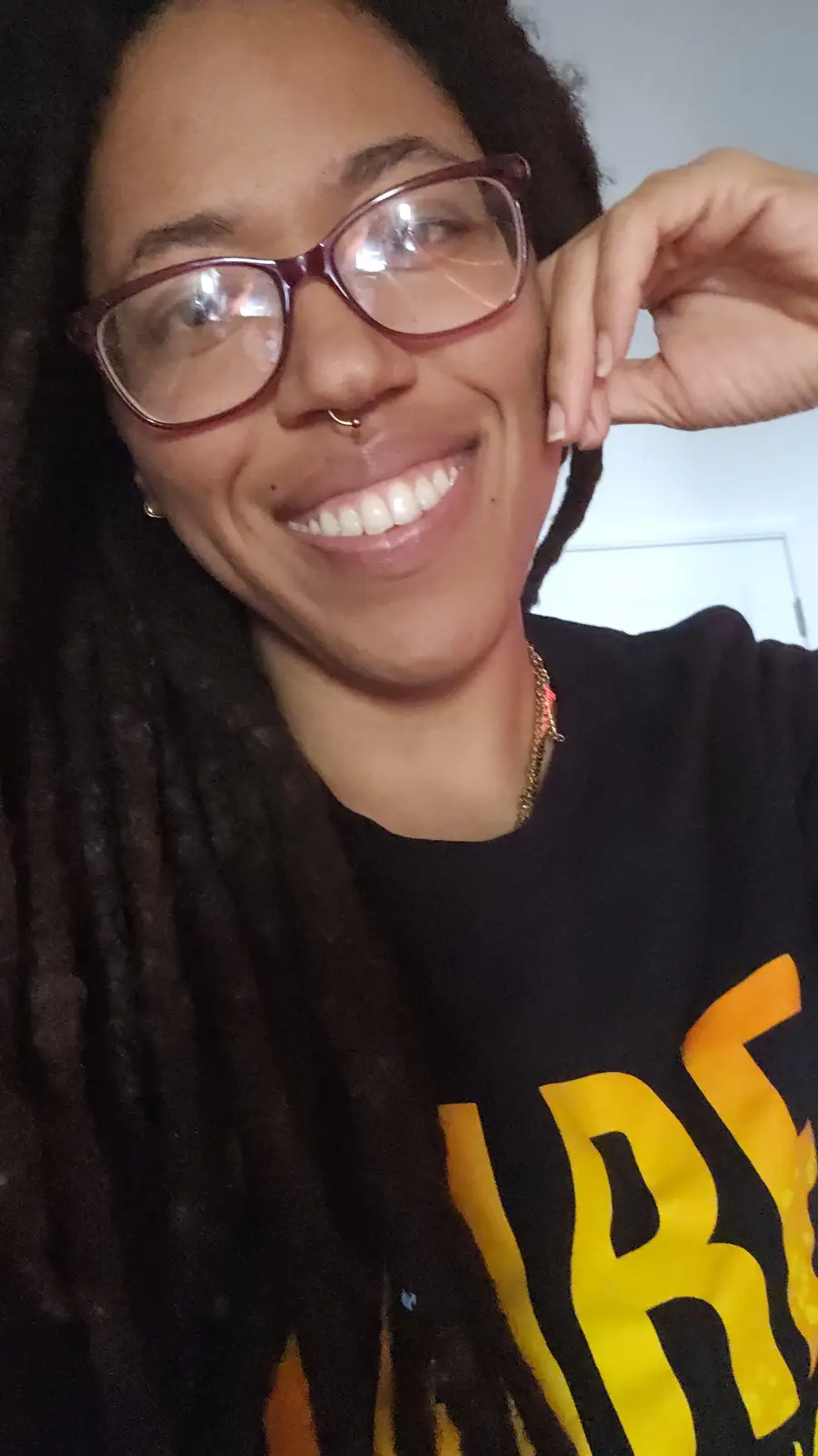 A smiling person with dark dreadlocks, wearing glasses and a nose ring, holds their hand to their ear as if listening…