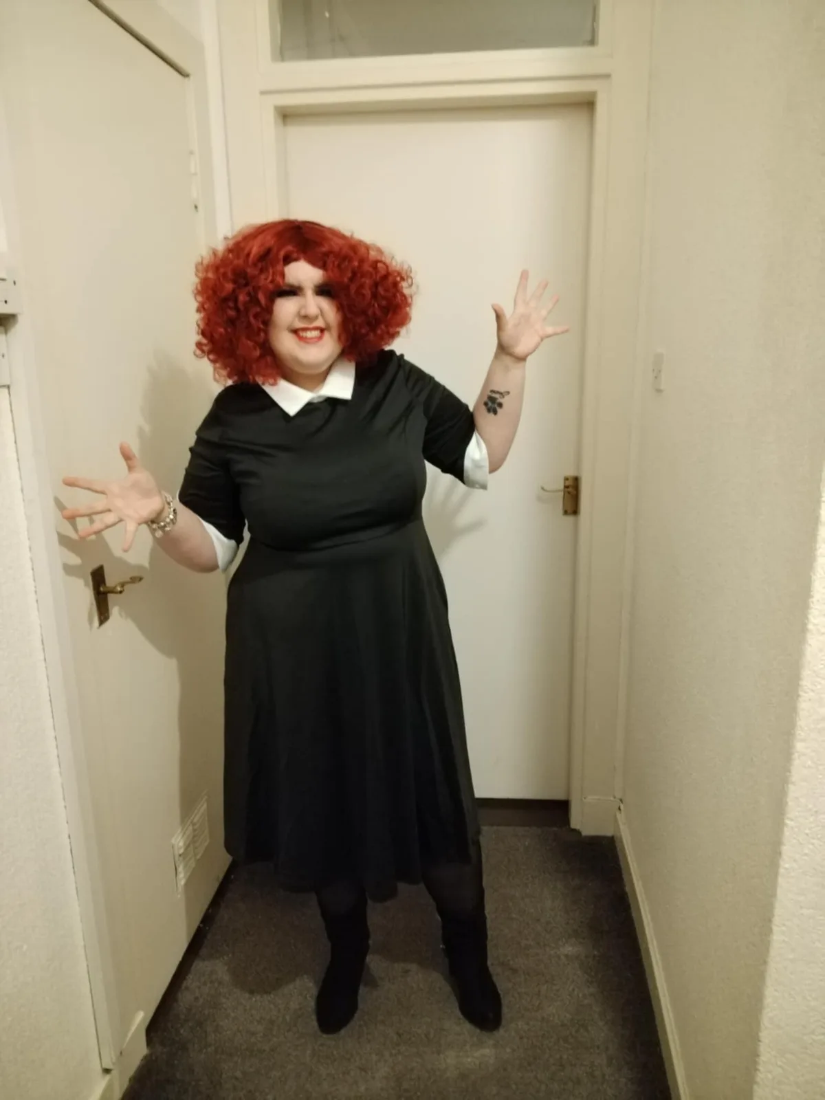 A person with a bright red curly wig and a black dress with a white collar stands in a hallway between two doors, arms…