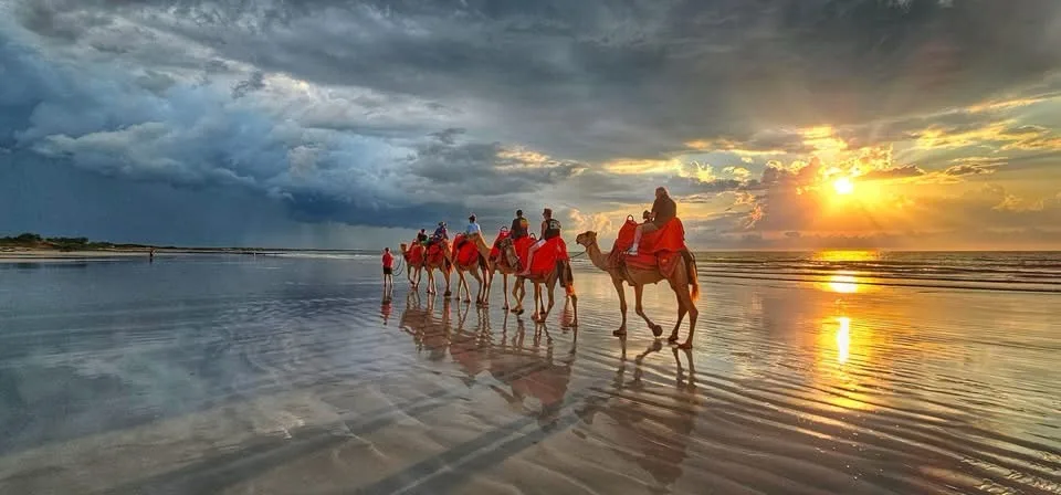 A line of people riding camels across a wet beach at sunset, with dramatic storm clouds and rain visible on the left side of…
