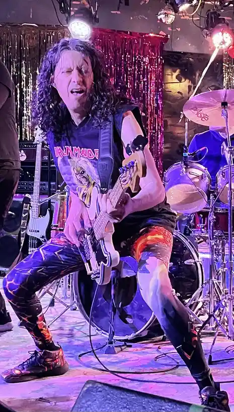 A man with long curly hair, wearing an Iron Maiden t-shirt and flame-patterned pants, plays a bass guitar on stage with…