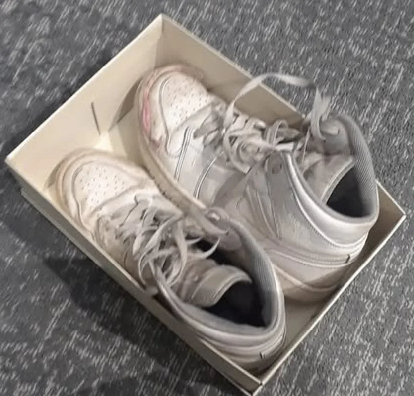 A pair of white high-top sneakers sits inside their original cardboard shoebox, which rests on a gray carpet. The shoes…
