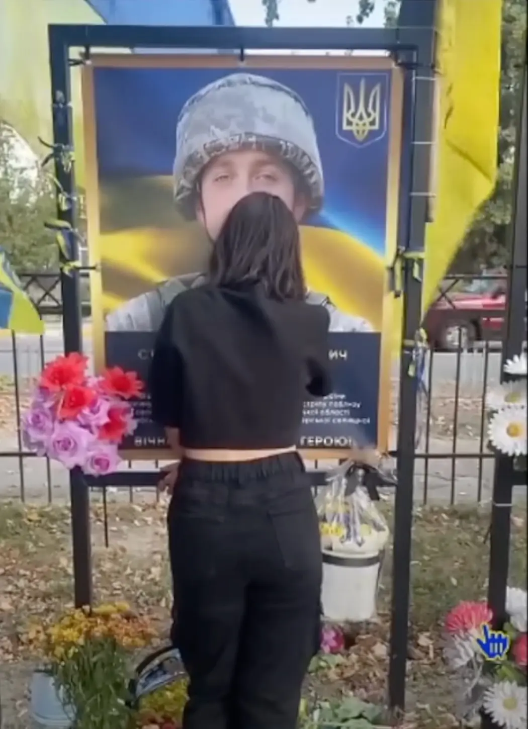 A woman in black clothing stands with her back to the camera, kissing a large poster of a Ukrainian soldier wearing a…