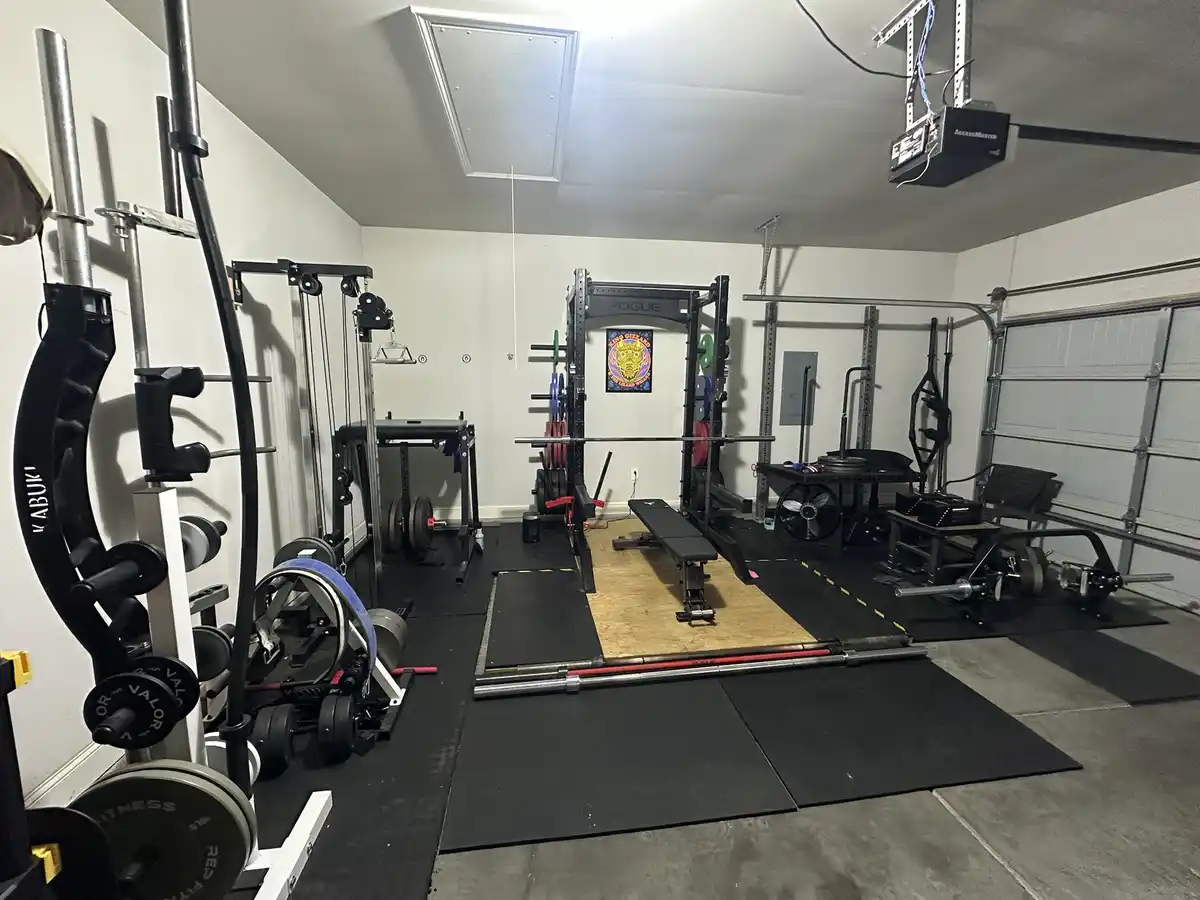 A well-equipped home gym in a garage, featuring a Rogue squat rack, weight plates, barbells, a cable machine, and various…