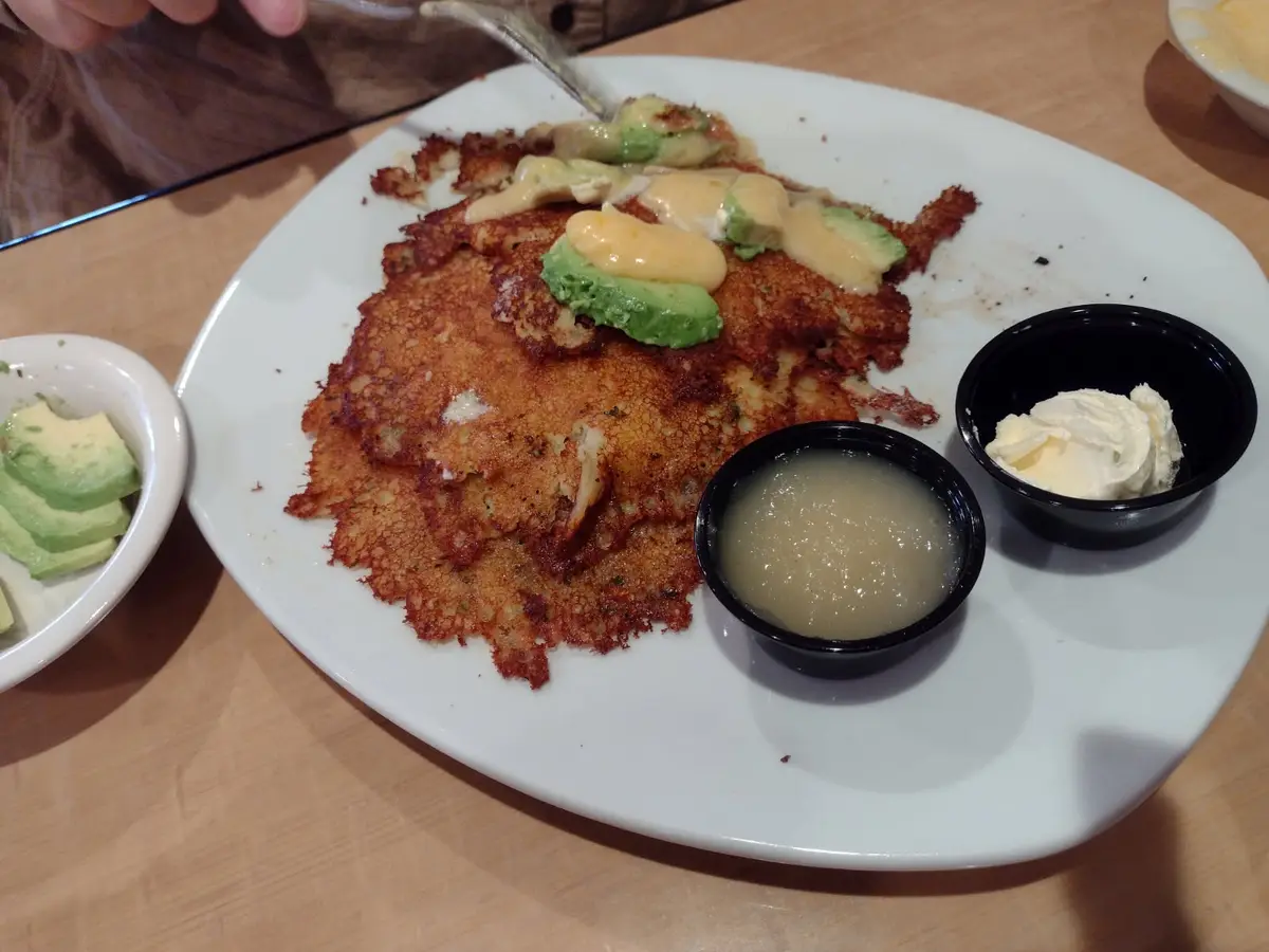 A close-up photo shows a large, golden-brown, crispy pancake-like dish on a white plate, topped with avocado slices and a…