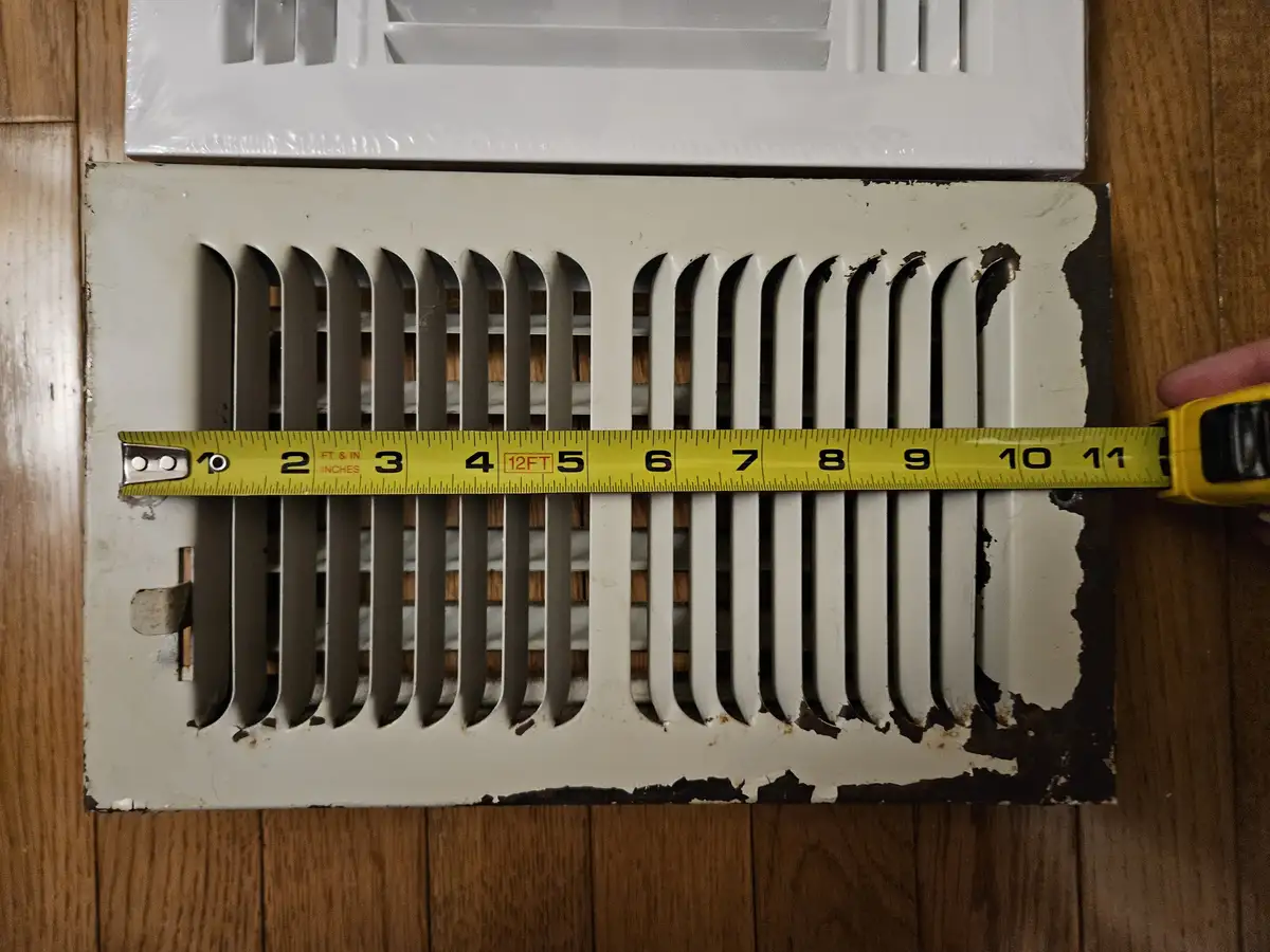 A yellow tape measure is stretched across a weathered, white metal floor vent, showing a measurement of 12FT. The vent has…