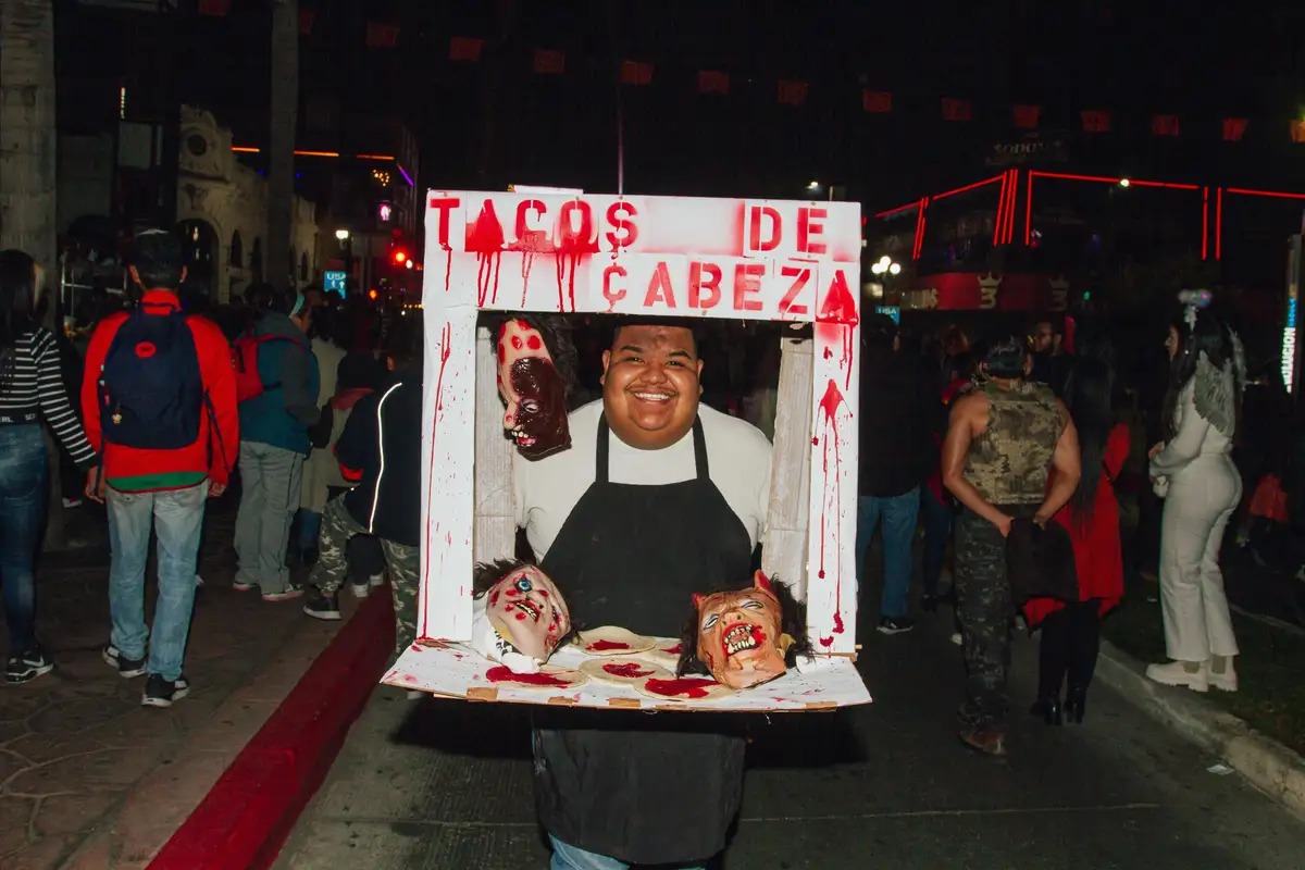A person in a black apron smiles while holding a makeshift food stand at night, decorated with fake bloody heads and red…