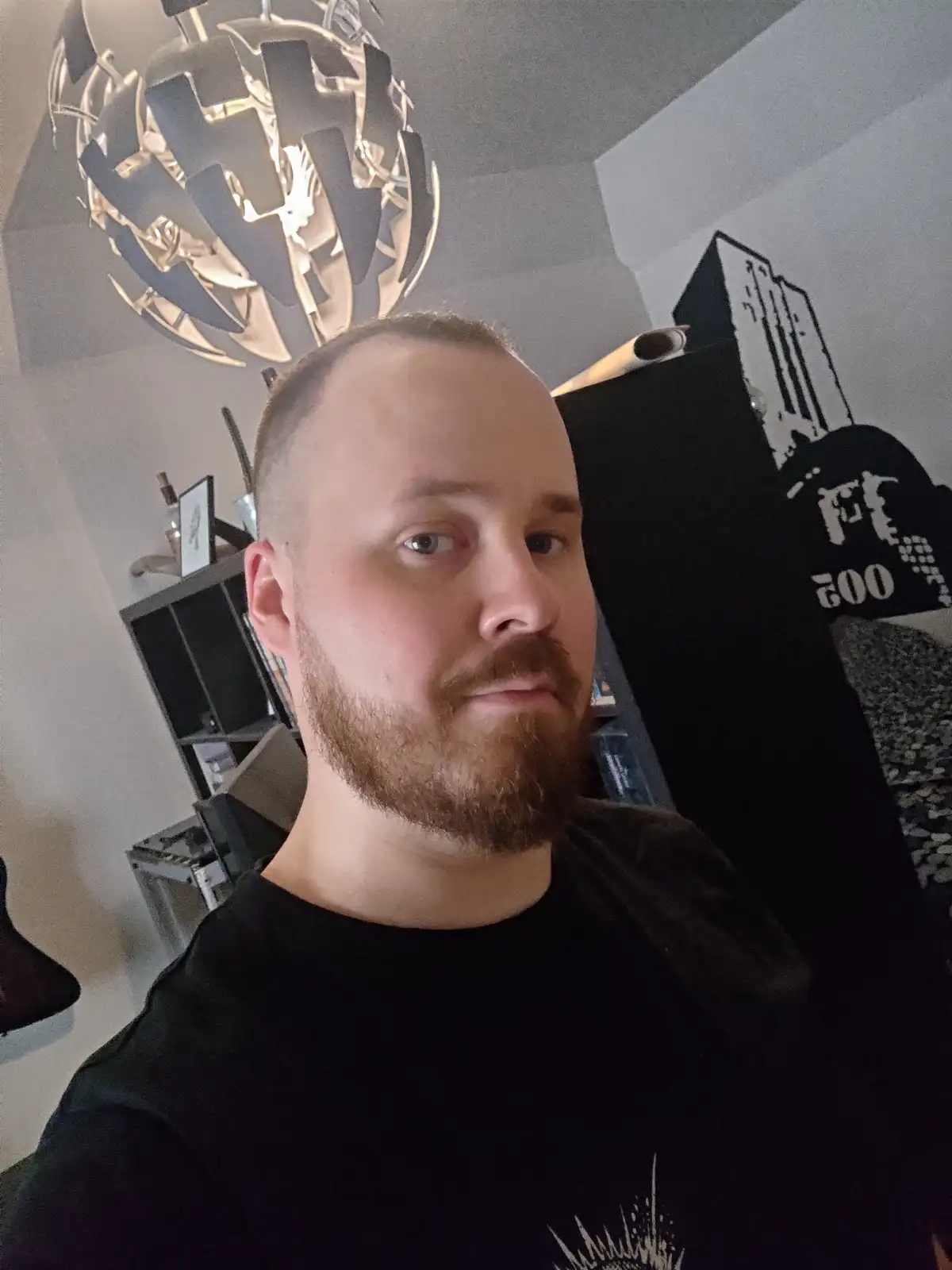 A man with a short haircut and beard stares directly at the camera with a neutral expression. Behind him, a large, modern…