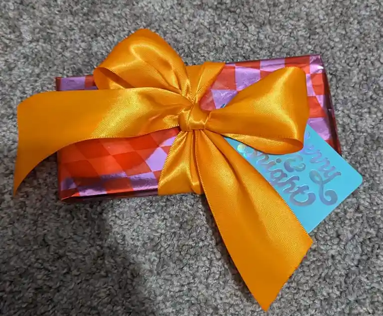 A small gift wrapped in shiny red and purple checkered paper, tied with a large, bright orange satin bow, rests on a gray…