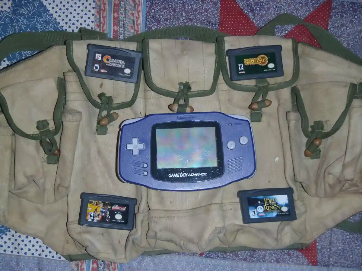 A blue Game Boy Advance sits in the center of a tan military-style utility vest with multiple pockets, each holding a game…