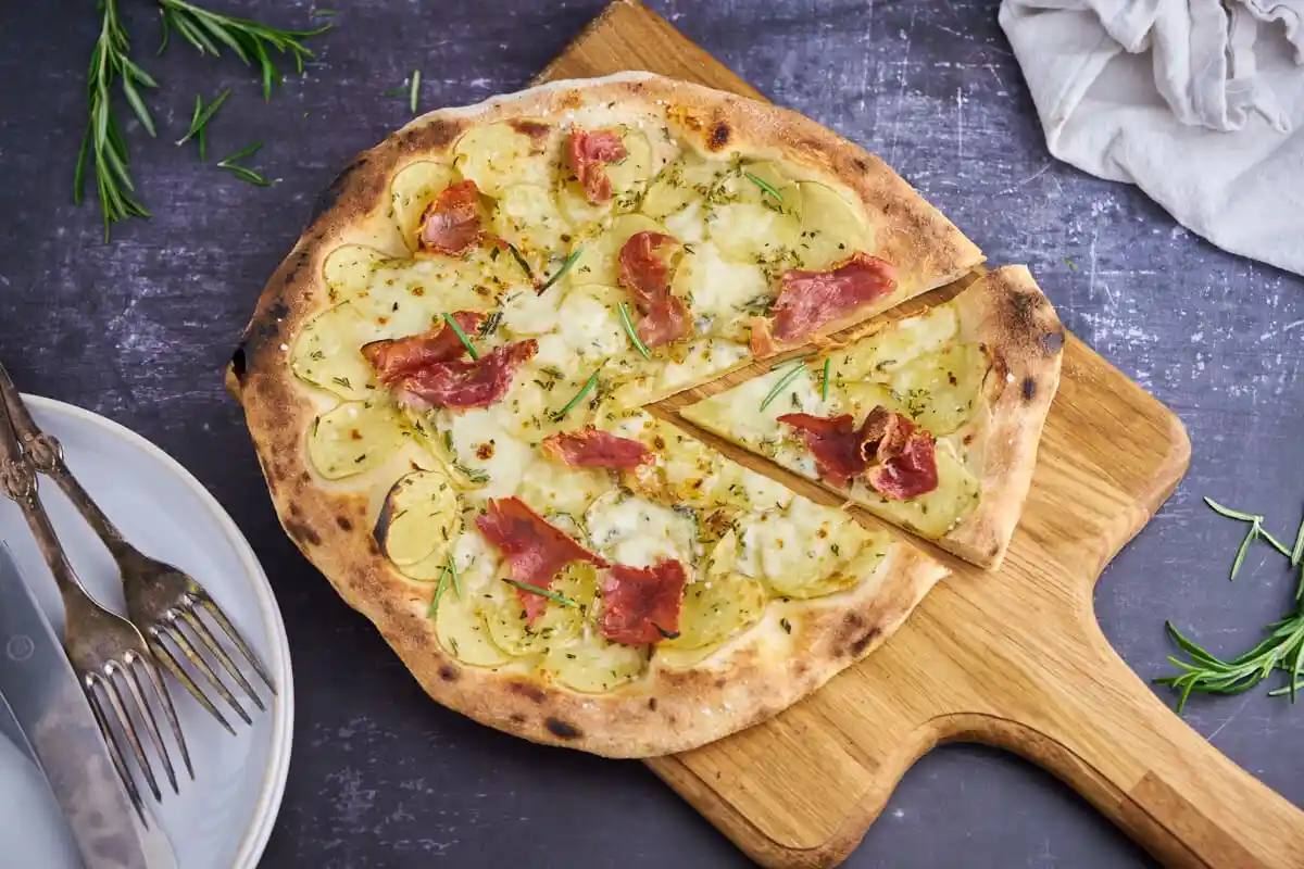 A rustic pizza with thin crust, topped with sliced potatoes, crispy bacon, melted cheese, and fresh rosemary, sits on a…