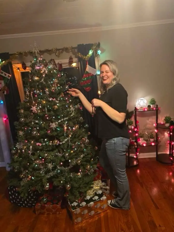 A woman with blonde hair, wearing a black t-shirt and jeans, stands beside a fully decorated Christmas tree in a warmly lit…